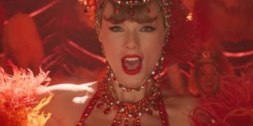 Taylor Swift in funkelndem roten Showgirl-Kostüm mit Federkopfschmuck und Kristallschmuck, Nahaufnahme auf einer Bühne mit warmem Rotlicht.