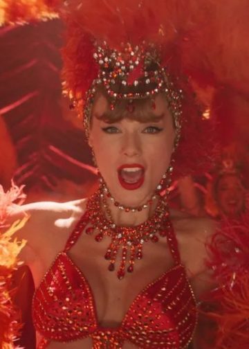 Taylor Swift in funkelndem roten Showgirl-Kostüm mit Federkopfschmuck und Kristallschmuck, Nahaufnahme auf einer Bühne mit warmem Rotlicht.