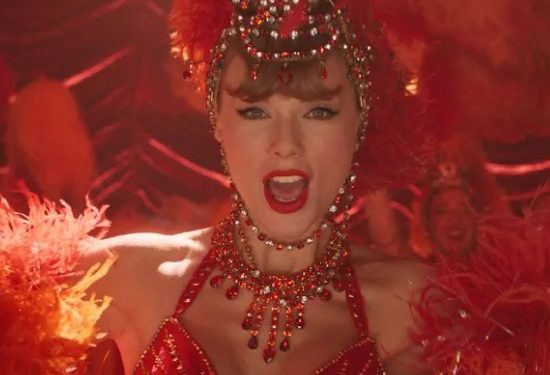 Taylor Swift in funkelndem roten Showgirl-Kostüm mit Federkopfschmuck und Kristallschmuck, Nahaufnahme auf einer Bühne mit warmem Rotlicht.