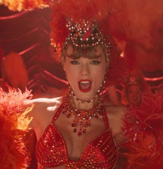 Taylor Swift in funkelndem roten Showgirl-Kostüm mit Federkopfschmuck und Kristallschmuck, Nahaufnahme auf einer Bühne mit warmem Rotlicht.